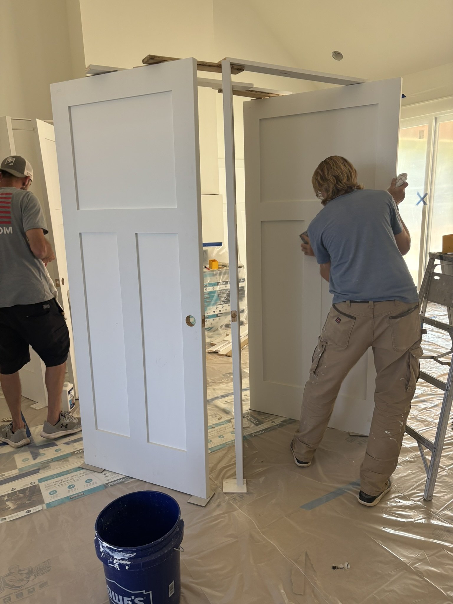 Two workers are installing white double doors inside a room under construction, with one worker on the left and another on the right, surrounded by construction materials and tools.