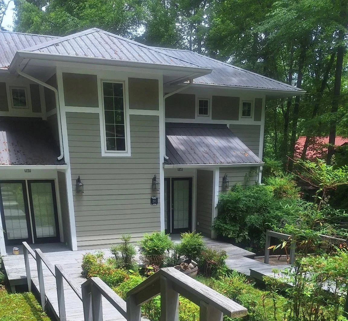 A two-story house with gray siding, white trim, and a metal roof surrounded by greenery and a wooden deck.