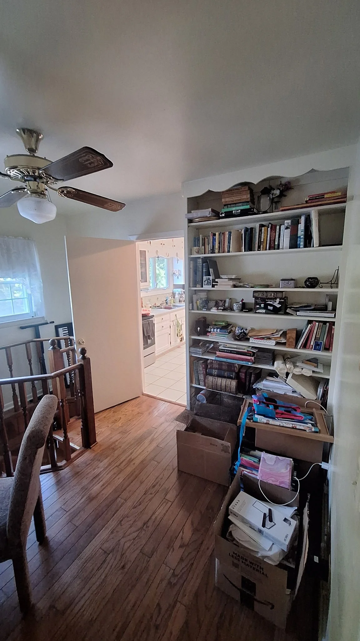 Living room with wooden floor, ceiling fan, white bookshelf filled with books and clutter, sitting area, adjacent kitchen with white cabinets and tiled floor, window with curtains, and staircase rail.