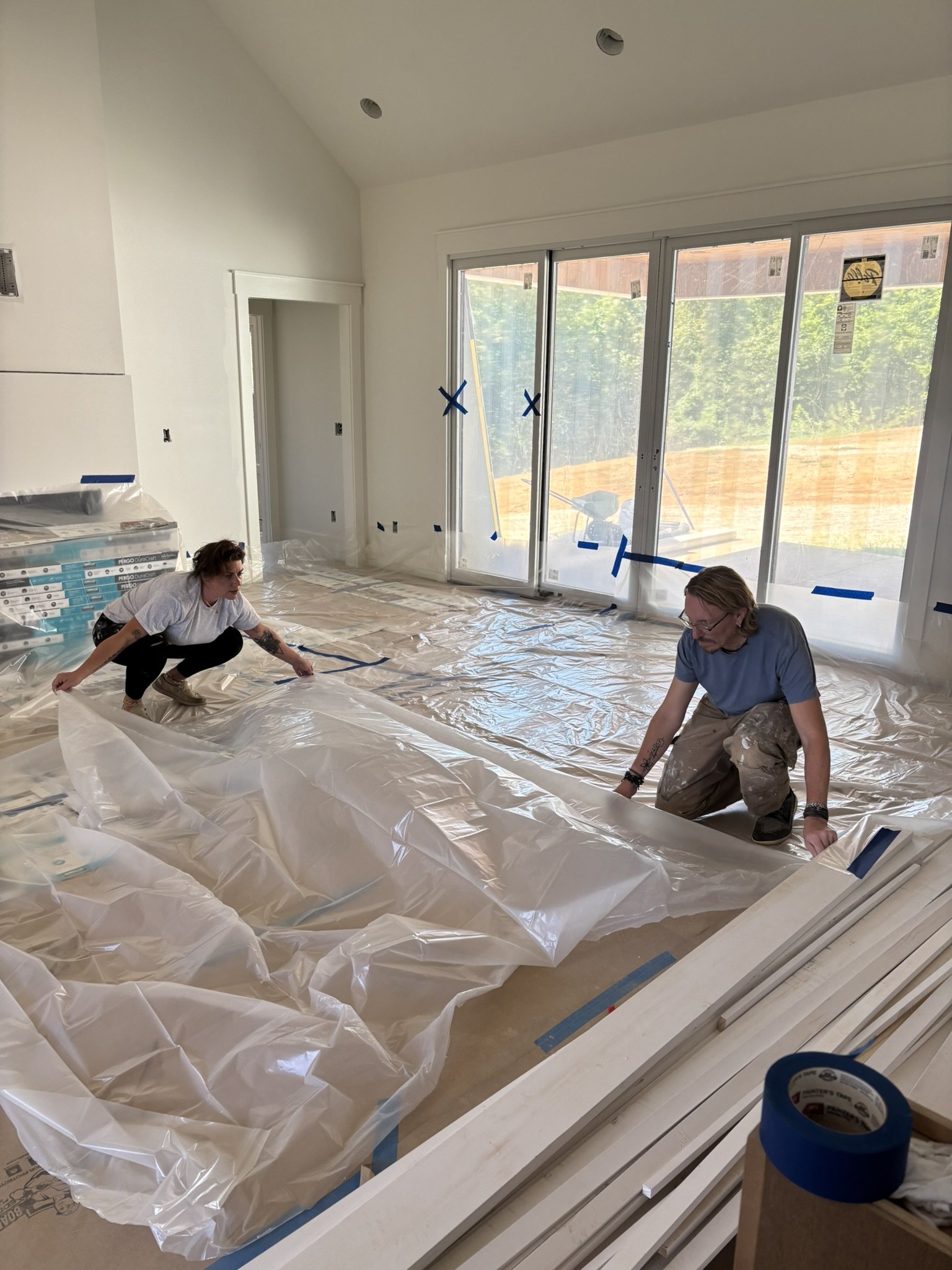 Two workers installing plastic sheeting on the floor of a room with large sliding glass doors during construction.