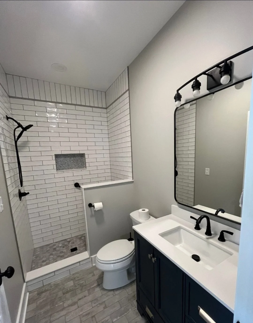 Modern bathroom with a walk-in shower, white subway tile walls, black fixtures, a vanity with a black faucet, and a large mirror, featuring gray wood-look tile flooring.
