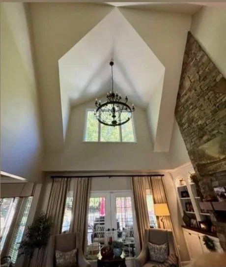 Living room with a high vaulted ceiling, large window, chandelier, curtains, and cozy furniture.