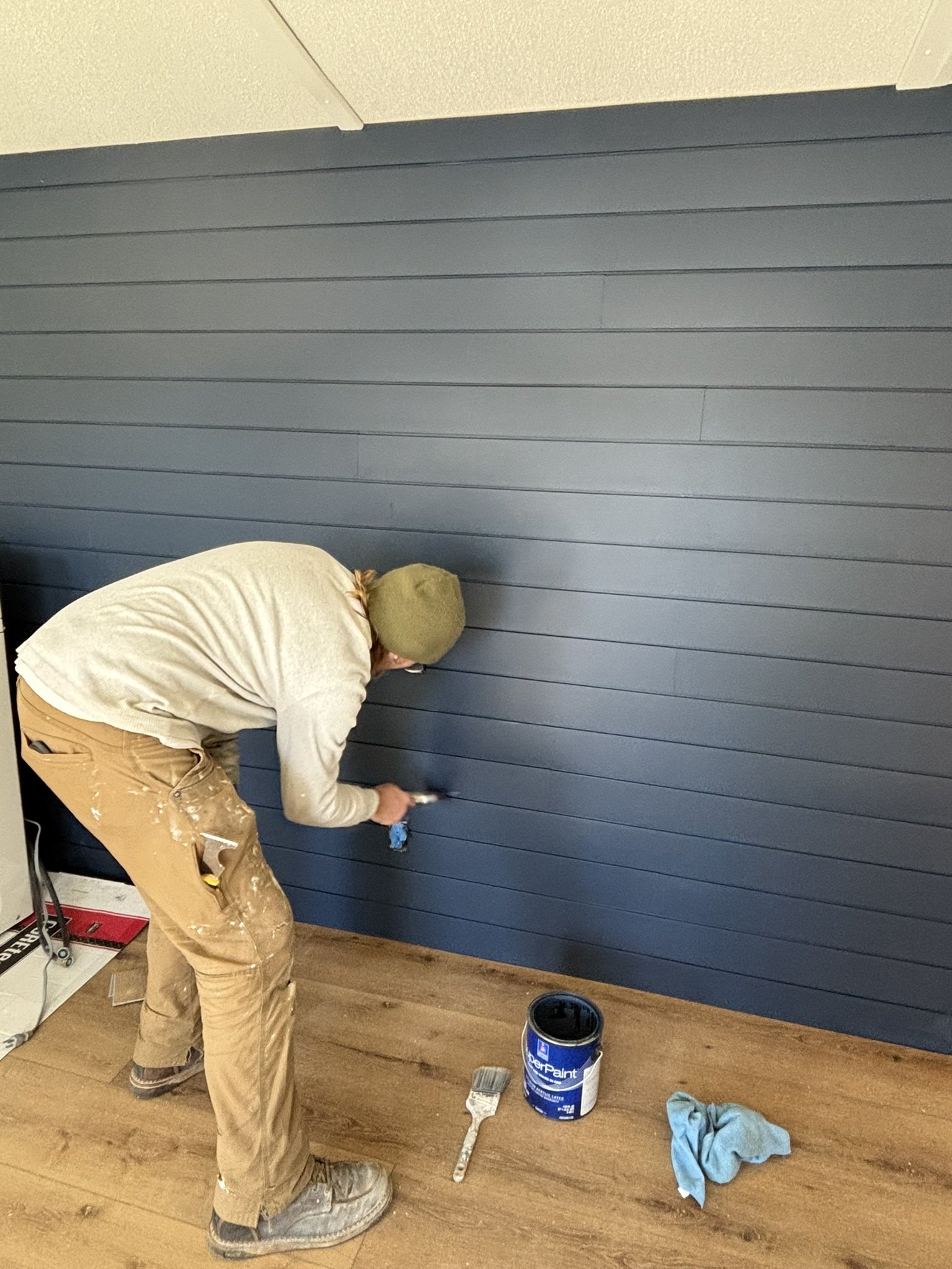 Person painting horizontal navy blue wood panel wall using a paintbrush, with an open can of navy paint and a paintbrush on the wooden floor.