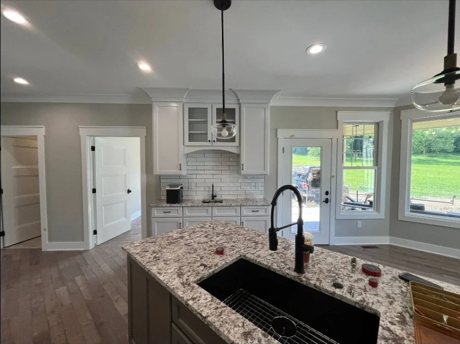 Modern kitchen with white cabinetry, granite countertops, and a kitchen island with a black sink and black faucet. Large windows and a glass door let in natural light, revealing a green lawn outside.