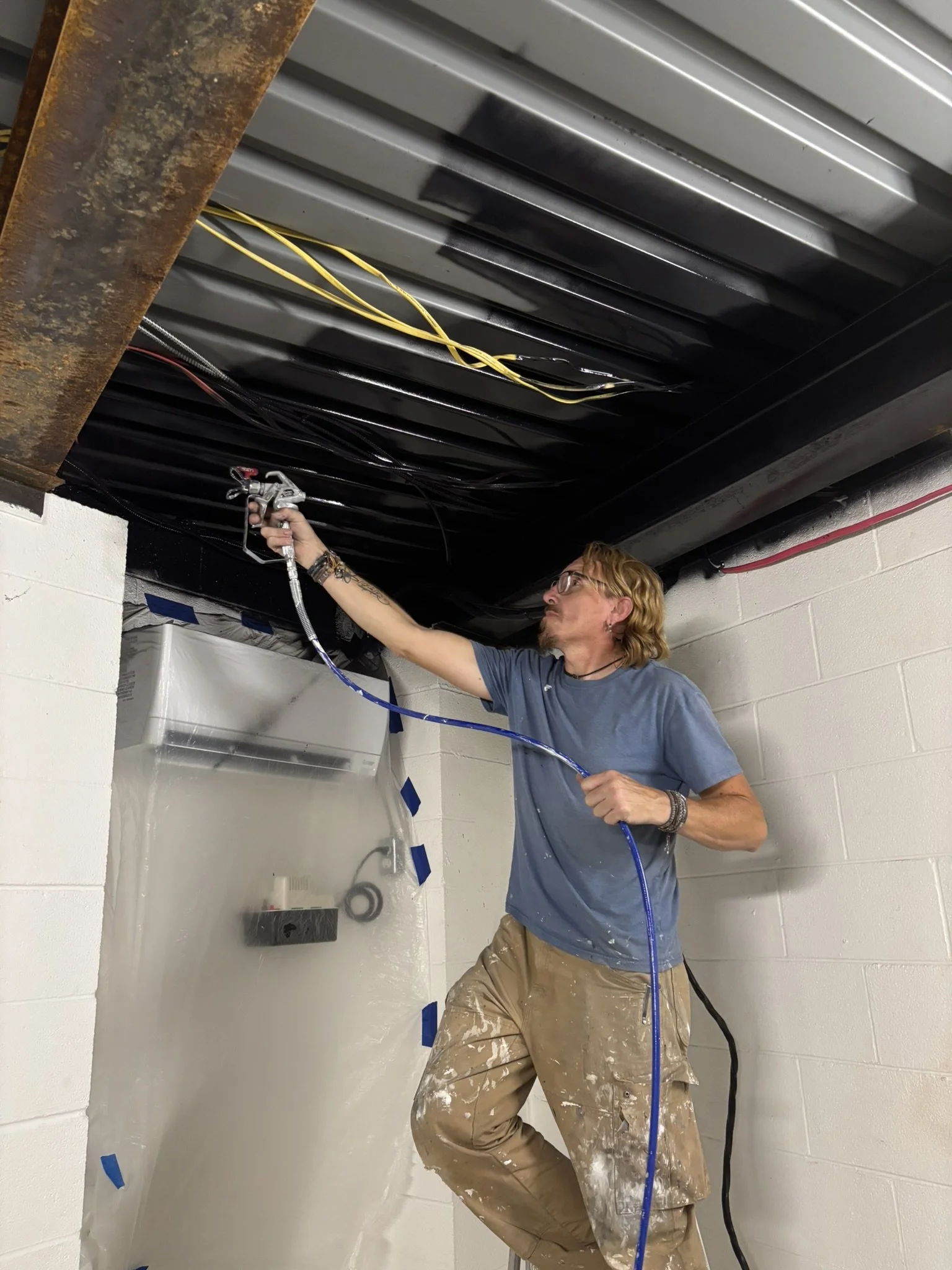 A man with blond hair and glasses spray-painting the underside of a black metal ceiling in an industrial or workshop setting.