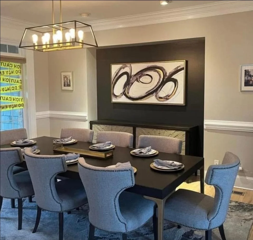 A dining room with a large, rectangular dining table set with plates, napkins, and silverware. Eight beige upholstered chairs are arranged around the table. A modern chandelier hangs above the table. There is a dark accent wall with abstract artwork above a sideboard. Windows on the left side are covered with yellow caution tape.