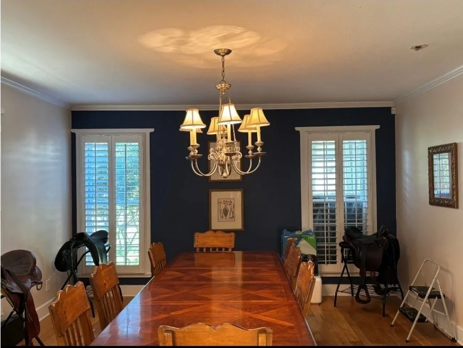 Dining room with a wooden table, six chairs, a chandelier with lampshades, two windows with blinds, and some photos and items on the walls.