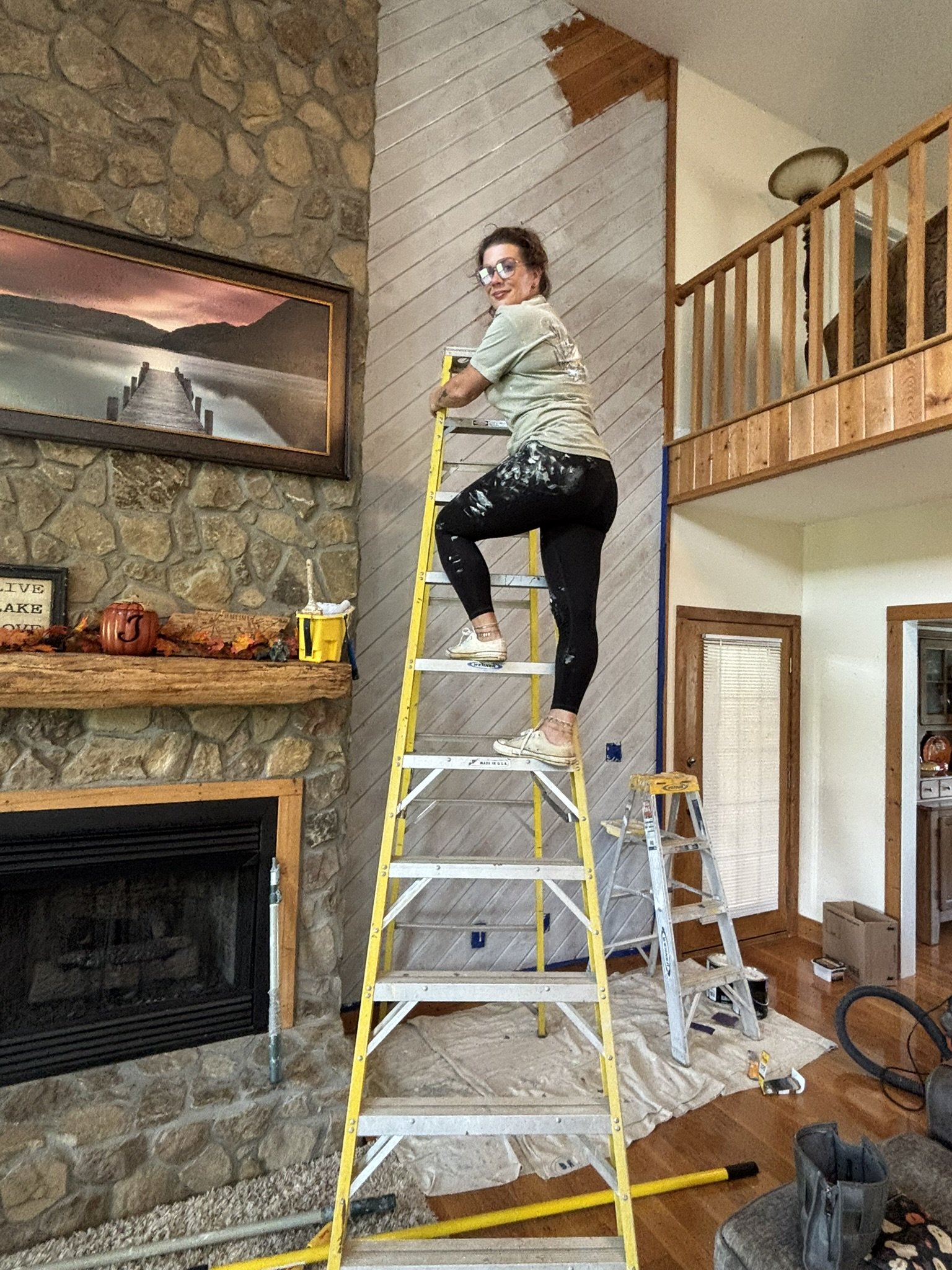 A woman is standing on a ladder painting a wall with diagonal wooden paneling in a living room, with a stone fireplace and a large framed landscape photograph on the wall.