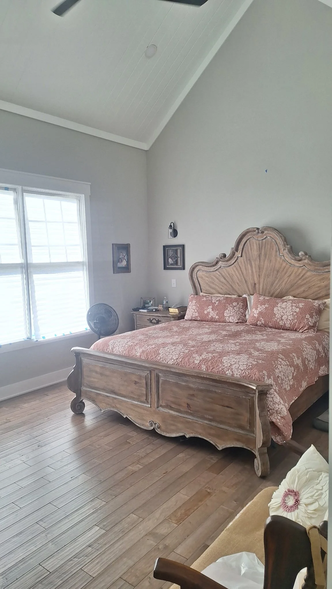 Bedroom with a wooden bed, pink floral bedding, window with blinds, nightstand with lamp, pictures on the wall, fan, and wooden floor.