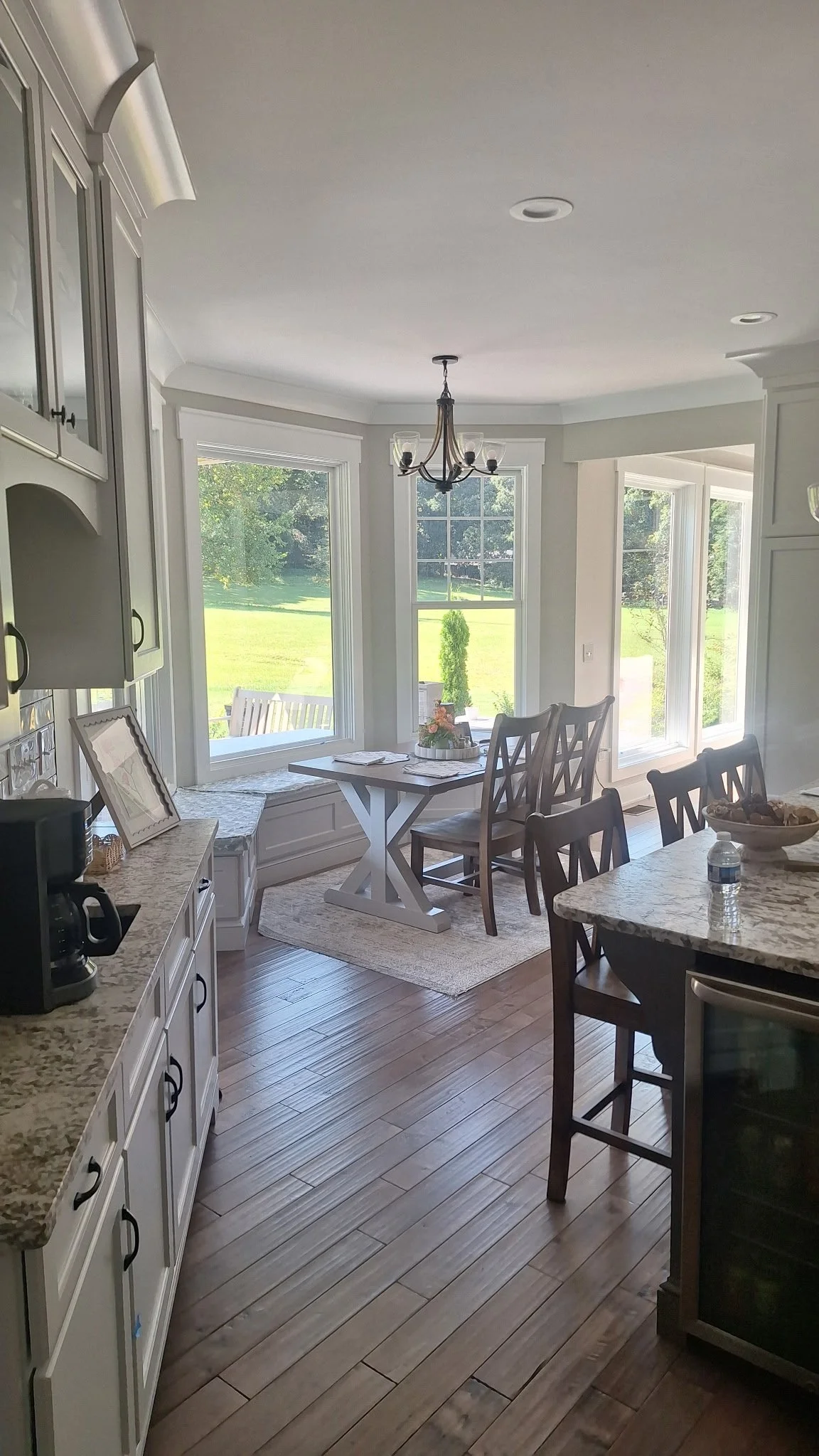 Bright kitchen and dining nook with large windows, wooden floors, white cabinetry, granite countertops, and a chandelier.