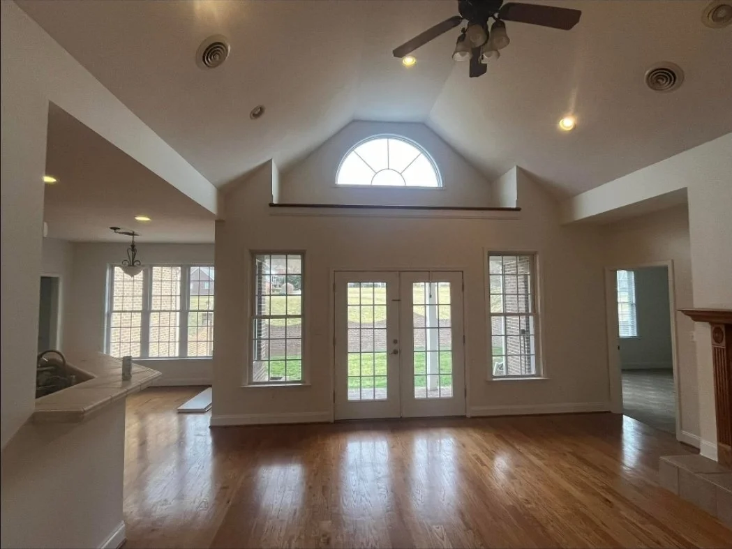 Interior of a spacious living room with high vaulted ceiling, large arched window above the door, double glass doors, and multiple side windows, with hardwood floors and minimal decor.
