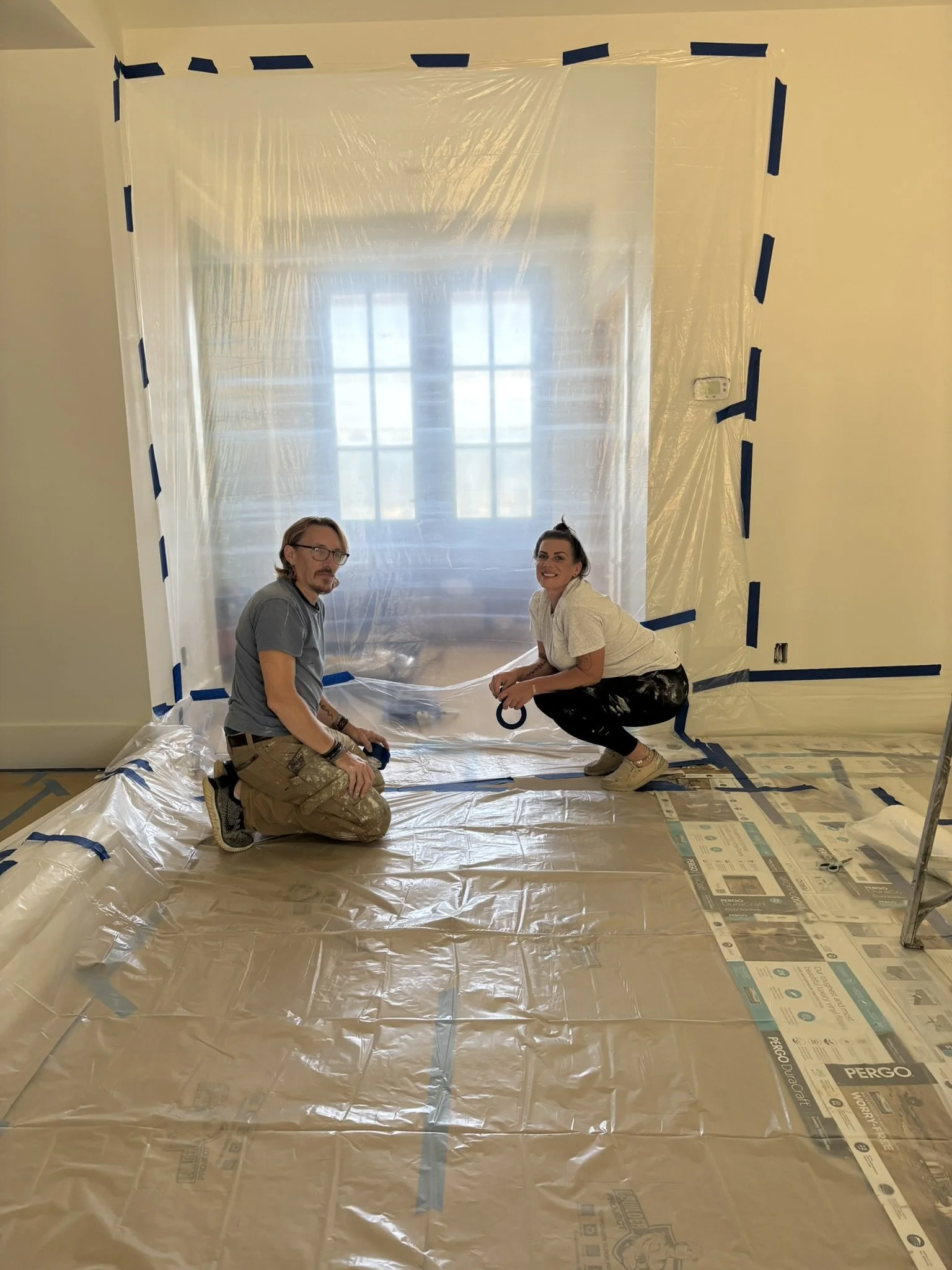 Two people kneeling on a plastic-covered floor in front of a wall covered with clear plastic and taped with blue painter's tape, working on a construction or renovation project in a room with a large window.