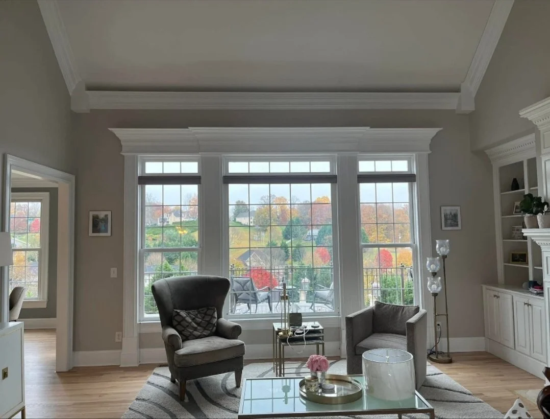 Living room with large bay window showing a colorful autumn landscape outside, two armchairs, a glass coffee table with a pink flower arrangement and decorative tray, a tall floor lamp, and built-in white shelving.