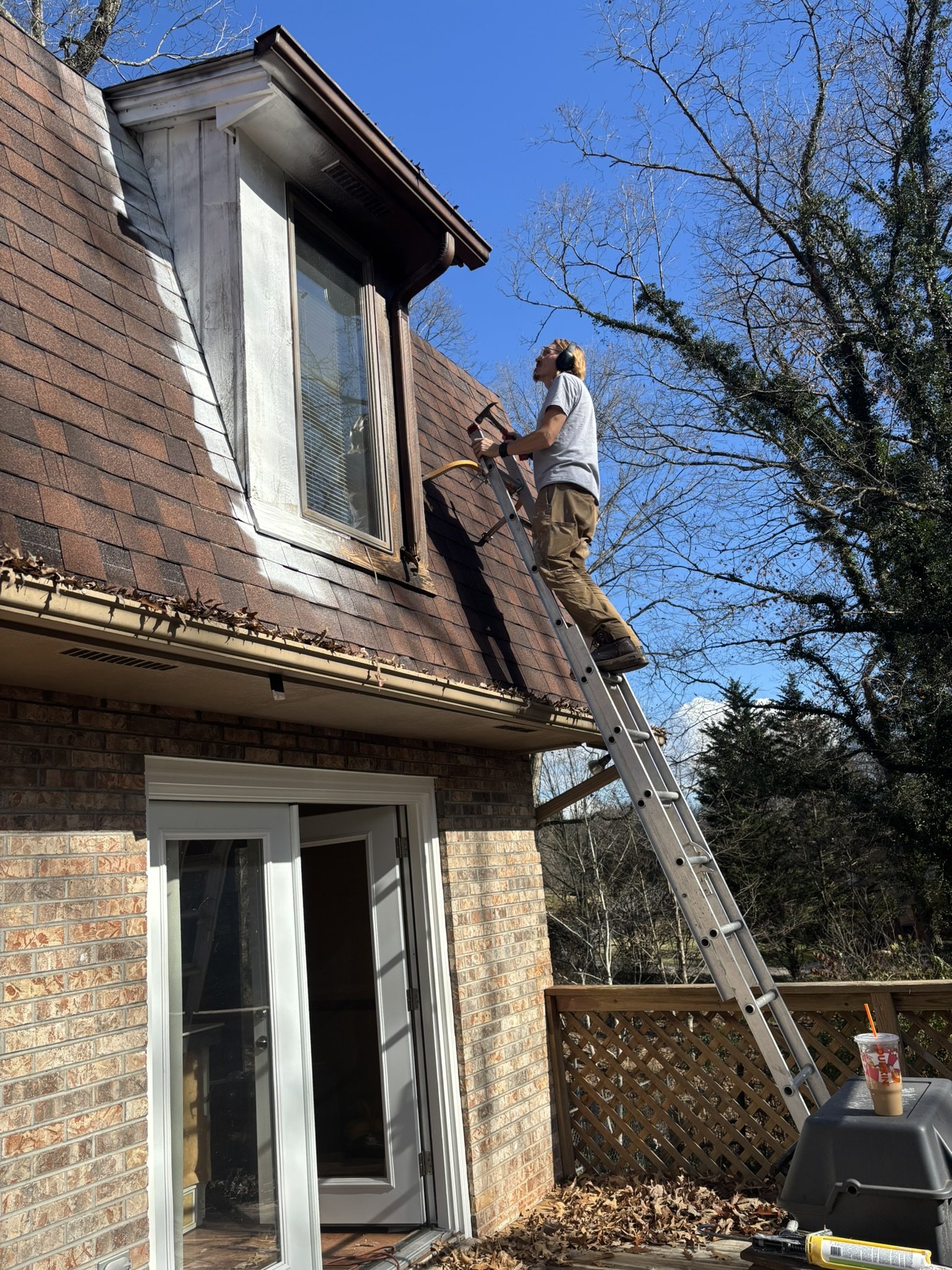 A person standing on a ladder cleaning or repairing a window on the roof of a house under a clear blue sky, with leafless trees in the background.