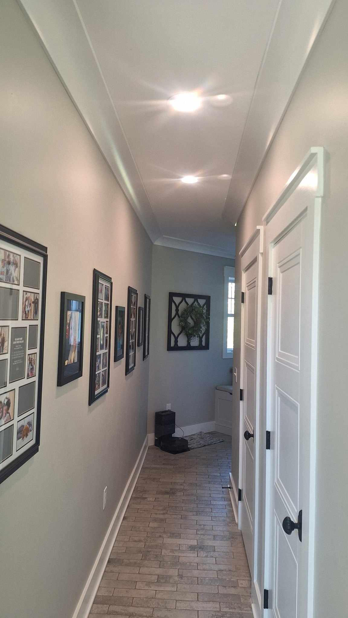 A house hallway with framed photos on the left wall, white double doors on the right, a small bench near the window at the end, and a decorative wall mirror with a plant in a black frame. The ceiling has two recessed lights.