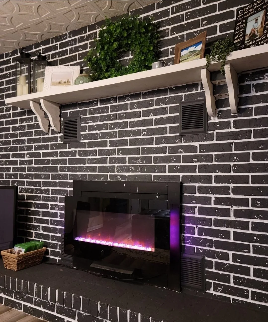 A black brick fireplace with a glowing electric fire insert and a dark mantel. Above the mantel is a white shelf holding decorative items including framed pictures, potted plants, and candles in lanterns.