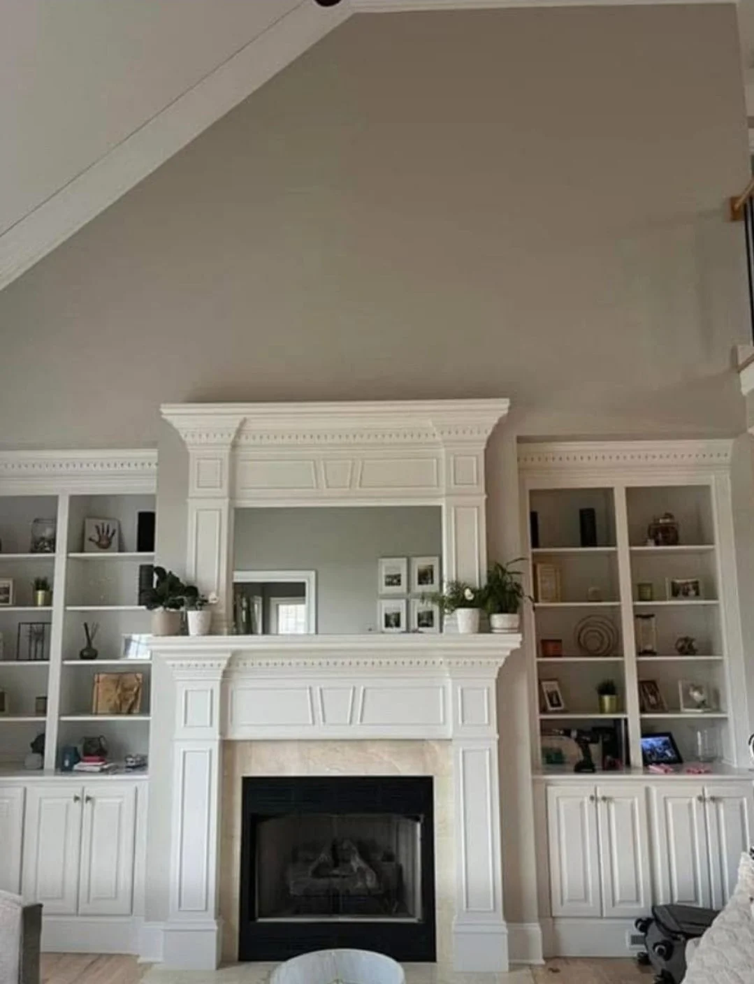 Interior view of a living room focused on a white fireplace with built-in shelves on either side, decorated with plants, picture frames, and decorative objects.