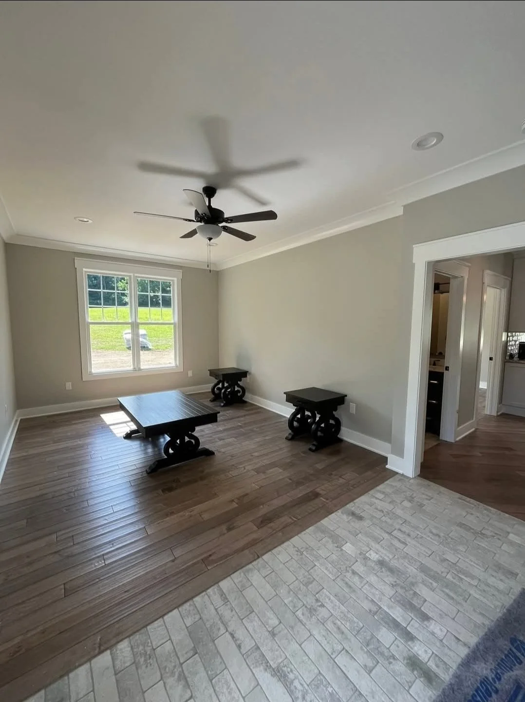 Empty living room with light-colored walls, a ceiling fan, a window with natural light, and a wooden floor transition to tile flooring near the entrance.