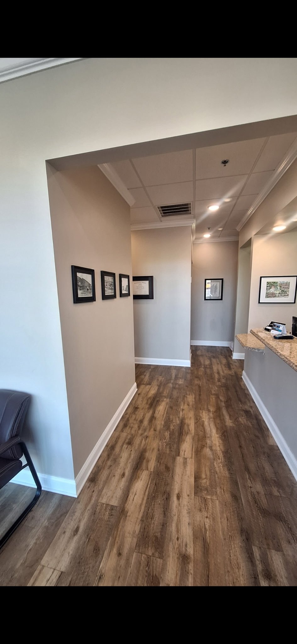 Interior view of an office or reception area with wood flooring, beige walls, and framed pictures on the walls, including a countertop with office supplies on the right side.