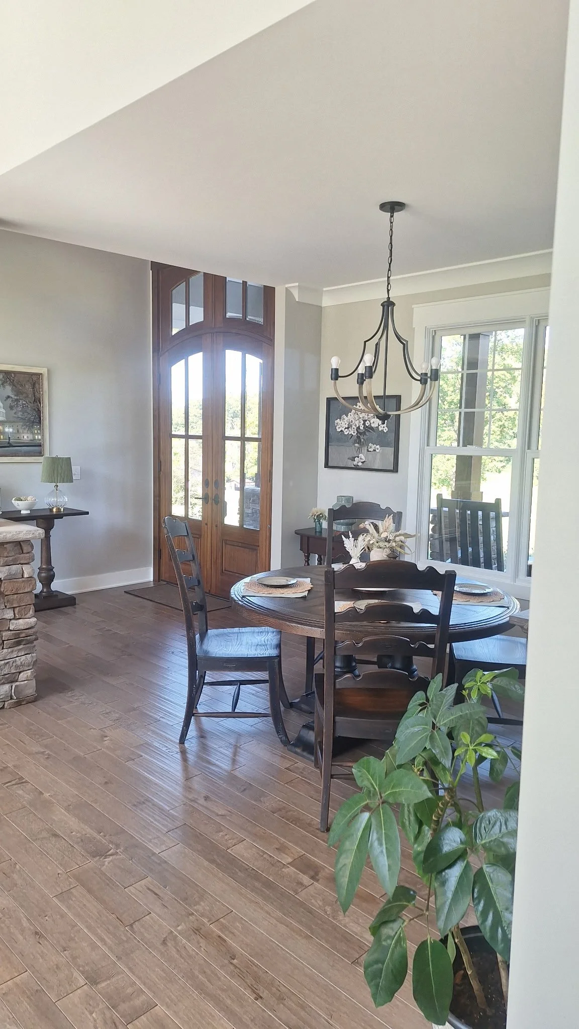 A dining room with a round wooden table set with plates and a centerpiece, surrounded by wooden chairs, a chandelier hanging above, a large window with a view of trees, a small side table with flowers and a framed picture, and a front door with glass