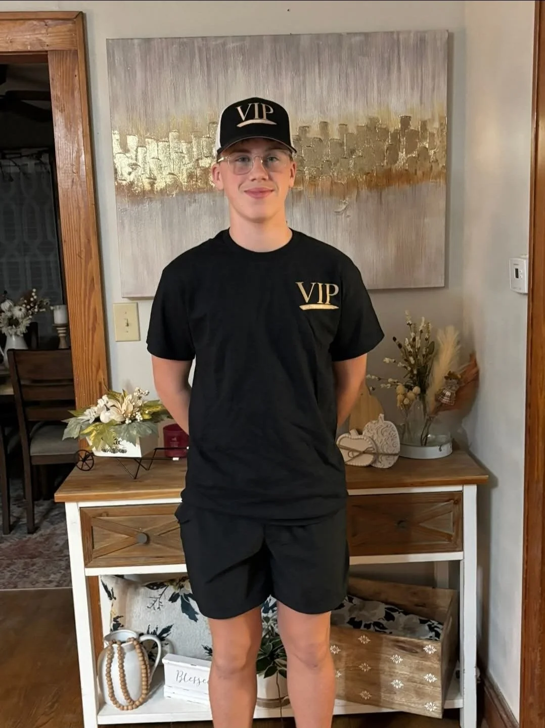 A young man wearing a black VIP hat and black T-shirt with VIP logo, standing indoors in front of a decorative wall art, with a console table decorated with flowers and plants behind him.