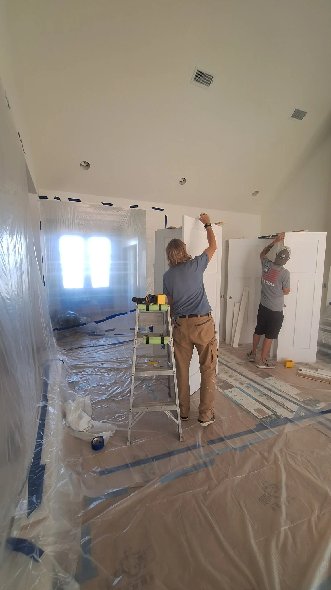Two people working on installing or painting built-in shelves or cabinets inside a room under construction or renovation, with plastic sheeting covering the floor and window for protection.