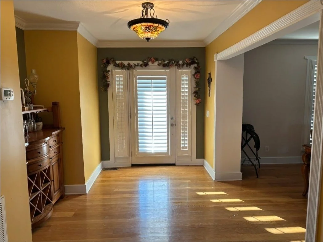 Dining room with yellow and green walls, wooden floor, and a decorative ceiling light. French doors with shutters lead outside, adorned with a floral garland. Part of a wooden china cabinet is visible on the left side, and a room with a chair and tab