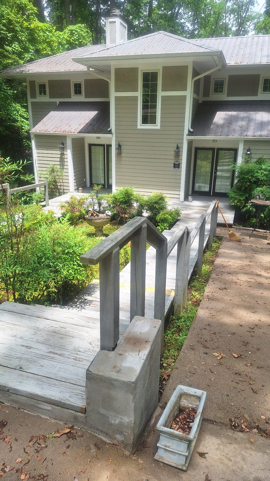 View of a two-story house with beige siding, metal roof, and a small front porch surrounded by lush green trees and shrubs.