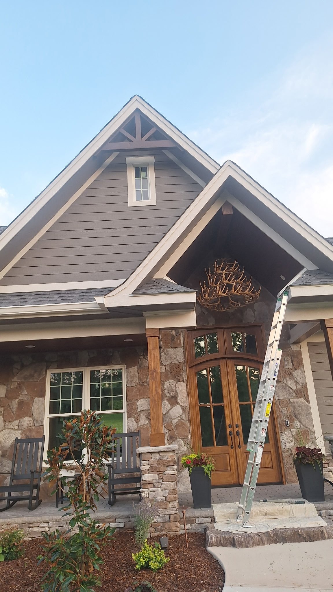 Front view of a house with a stone and wood exterior, a front porch with seating, potted plants, and a ladder leaning against the house near the entrance. The house has a pitched roof and decorative antlers above the front door.