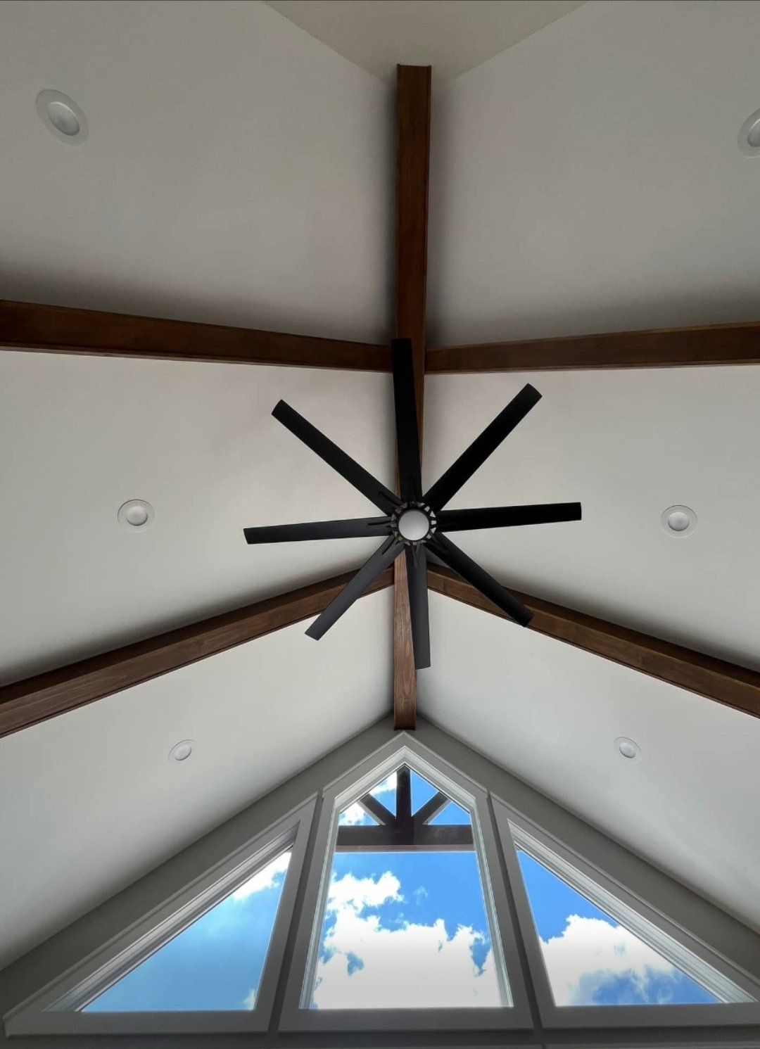 View of a modern interior ceiling with a black ceiling fan, wooden beams, and a triangular glass window showing a blue sky with white clouds.