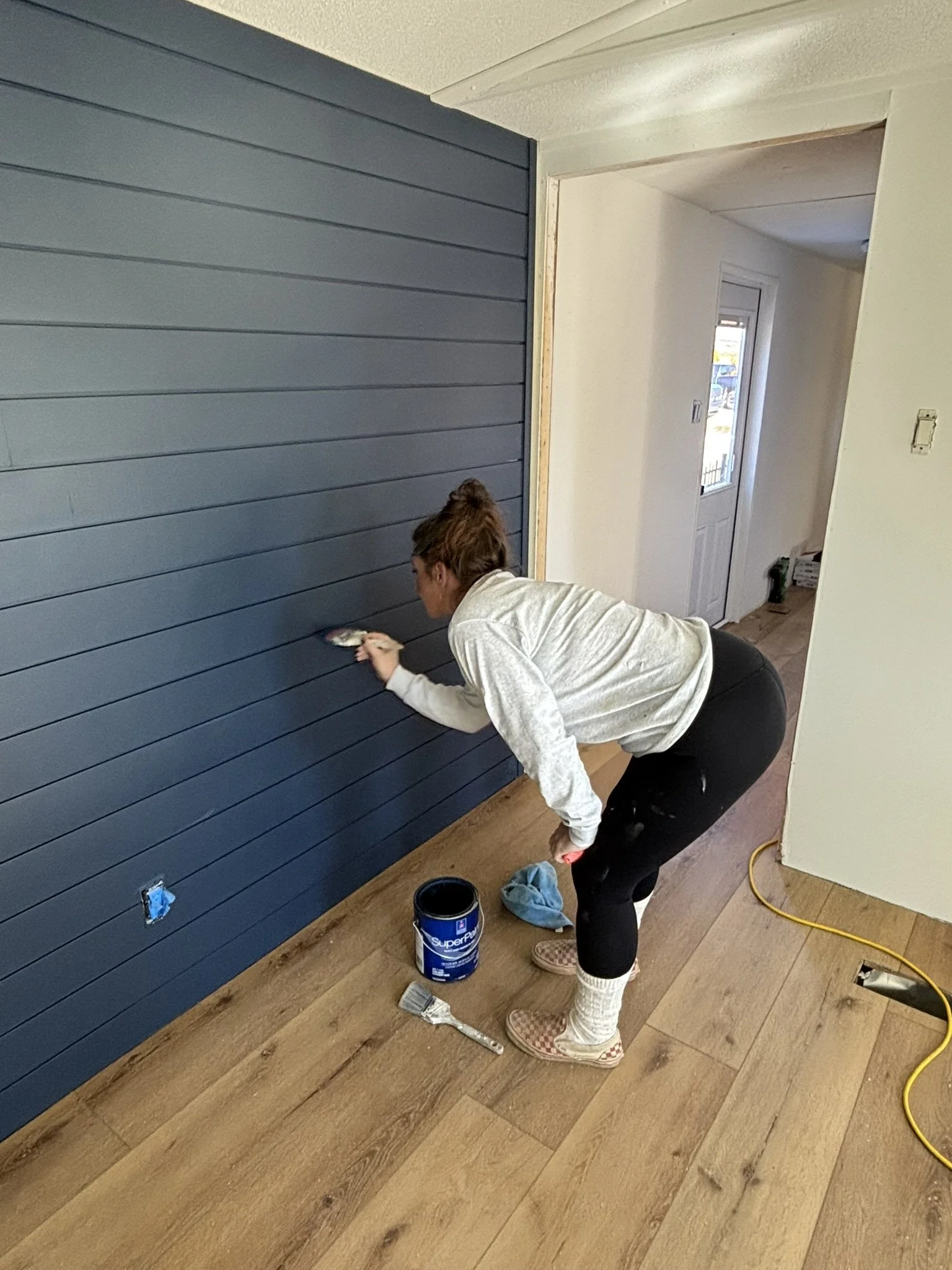 A woman painting a navy blue wooden wall in a house, standing on the floor with paintbrush in hand, wearing pajamas, slippers, and socks, with paint supplies and a yellow extension cord on the floor.