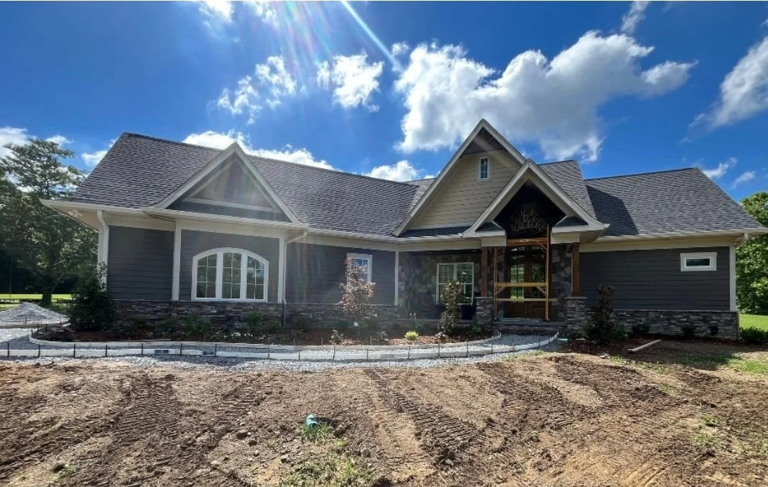A newly constructed house with a dark gray exterior, large front windows, and a gabled roof, set against a bright blue sky with white clouds, with landscaped front yard under development.