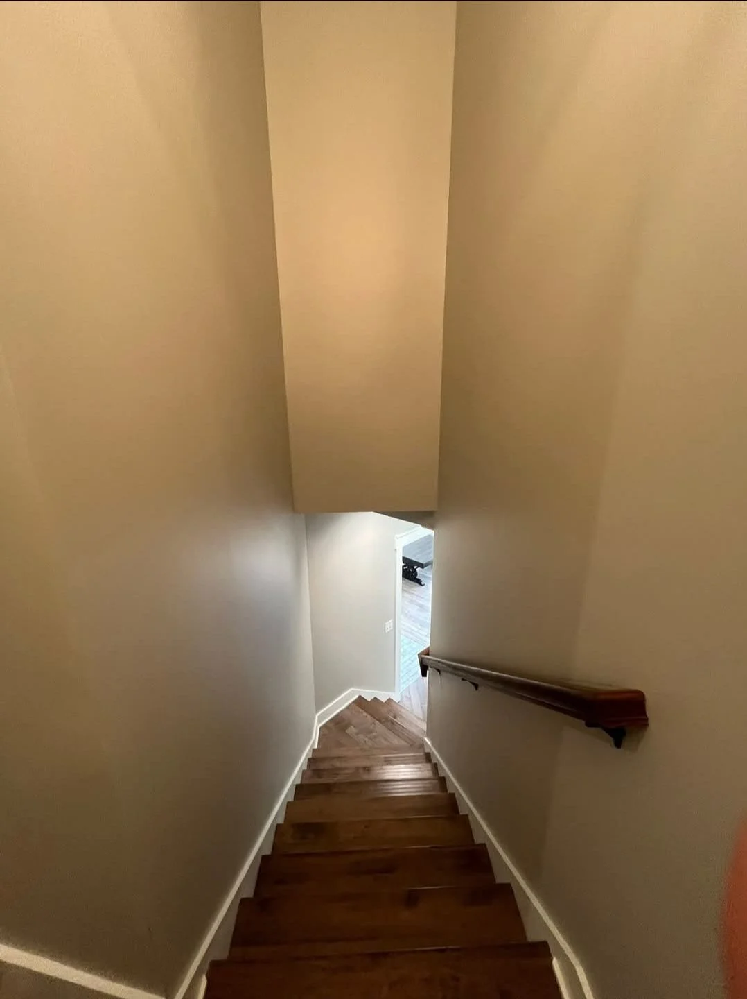 Interior view of a staircase with wooden steps, beige walls, and a wooden handrail on the right side, leading down to a room with a small portion of a floor and furniture visible.