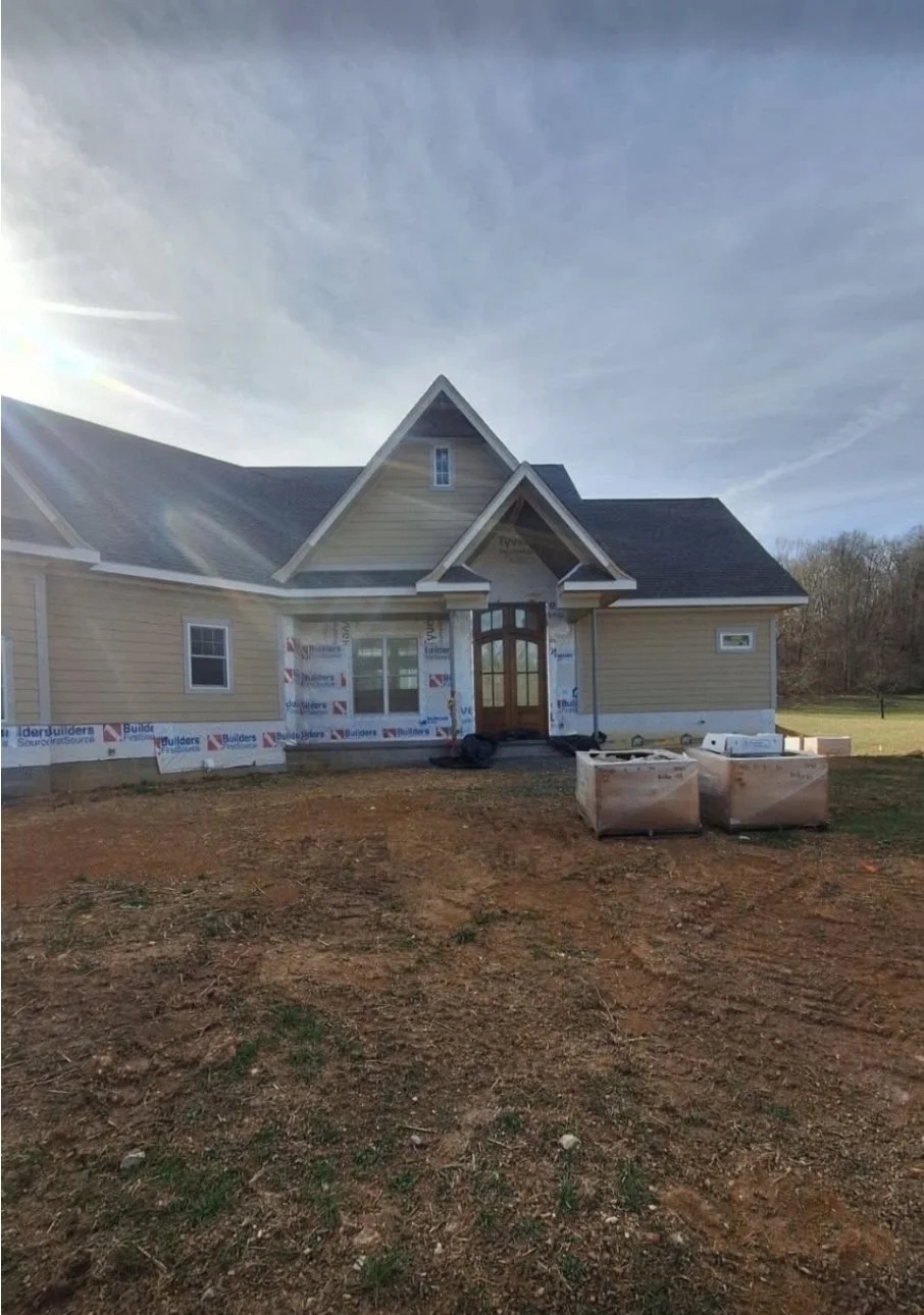 Newly constructed house with beige siding and a dark roof, front door with glass panels, and construction materials on the front yard, under a partly cloudy sky.