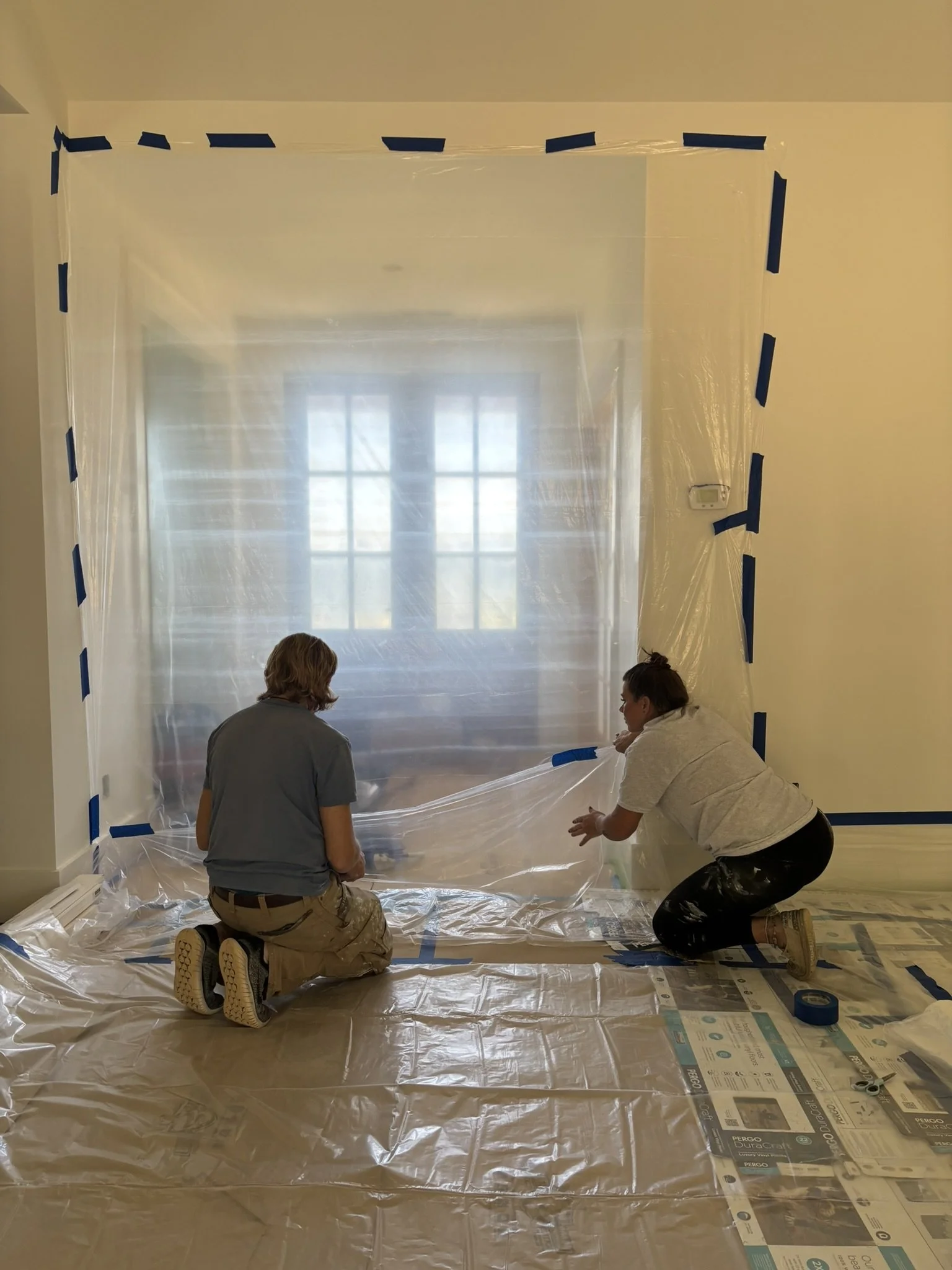 Two people kneeling on the floor, installing a plastic sheet around a window opening during a home renovation.