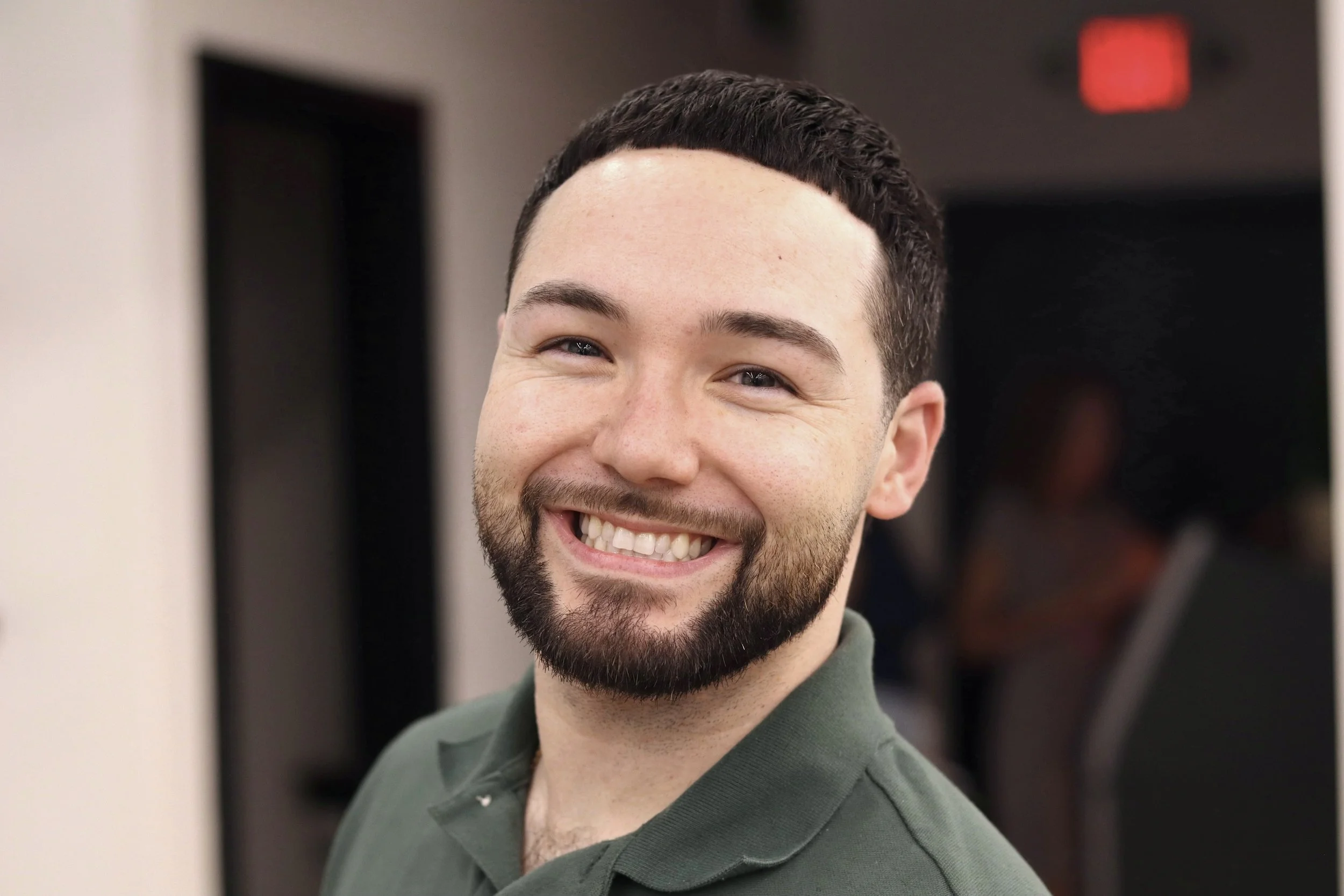 A smiling man with short dark hair and a beard, wearing a green collared shirt, standing indoors with a blurred background.