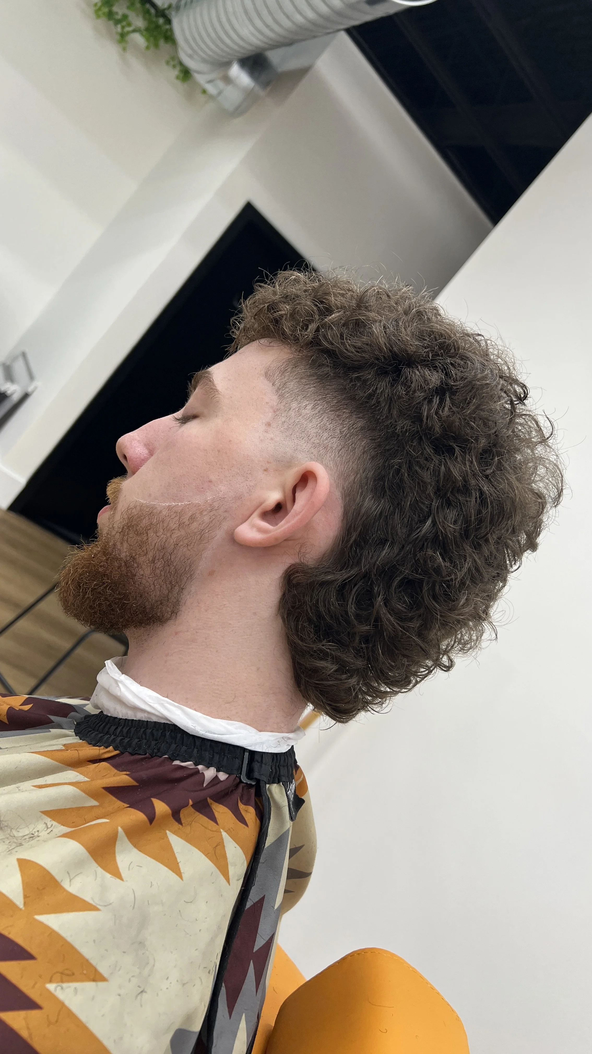 Side view of a young man with curly hair and a beard getting a haircut in a Barbershop, he has a curly modern mullet with a taper on the side 
