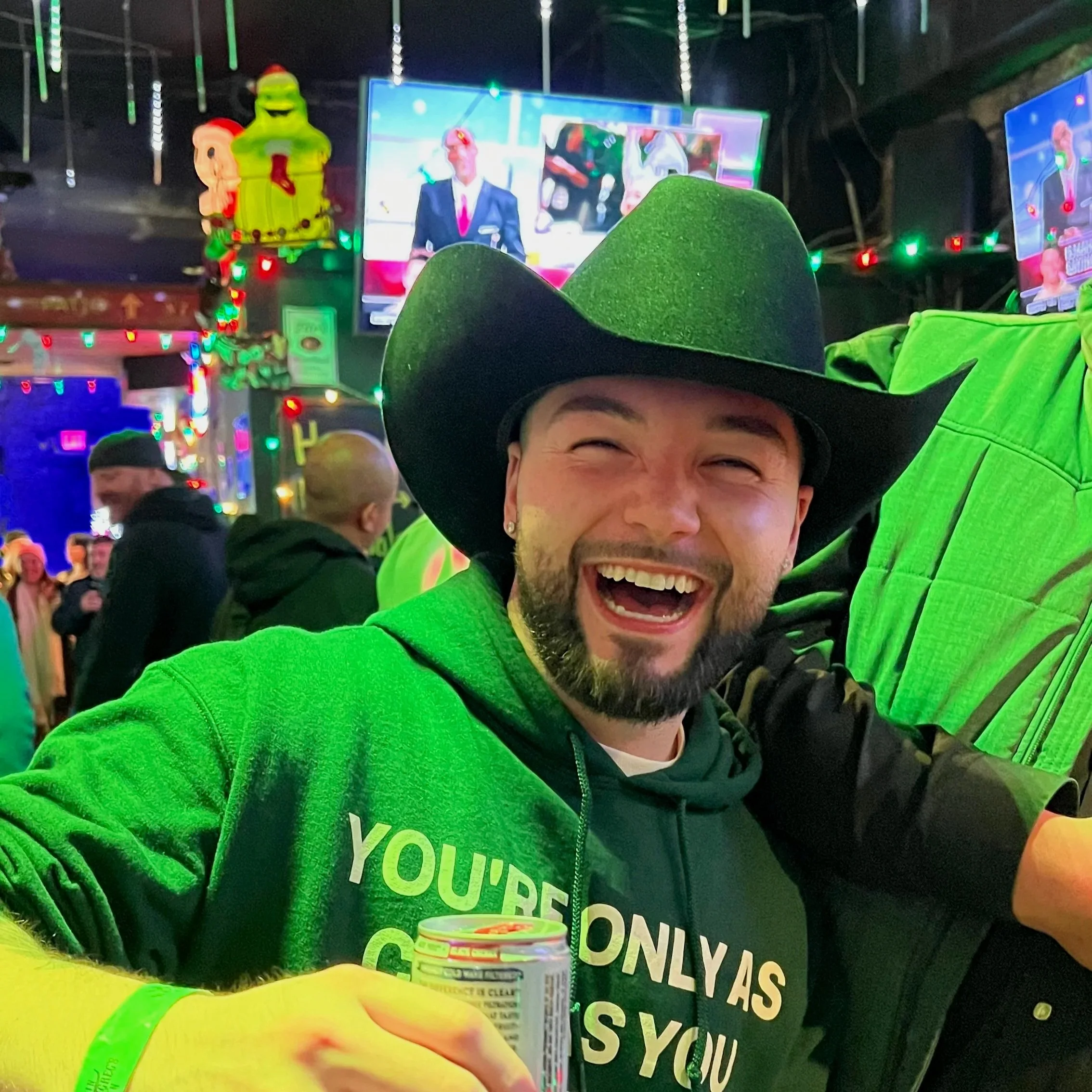 Smiling man wearing a black cowboy hat and bright green jacket taking a selfie at a brightly decorated party or event with Christmas lights and colorful displays in the background.