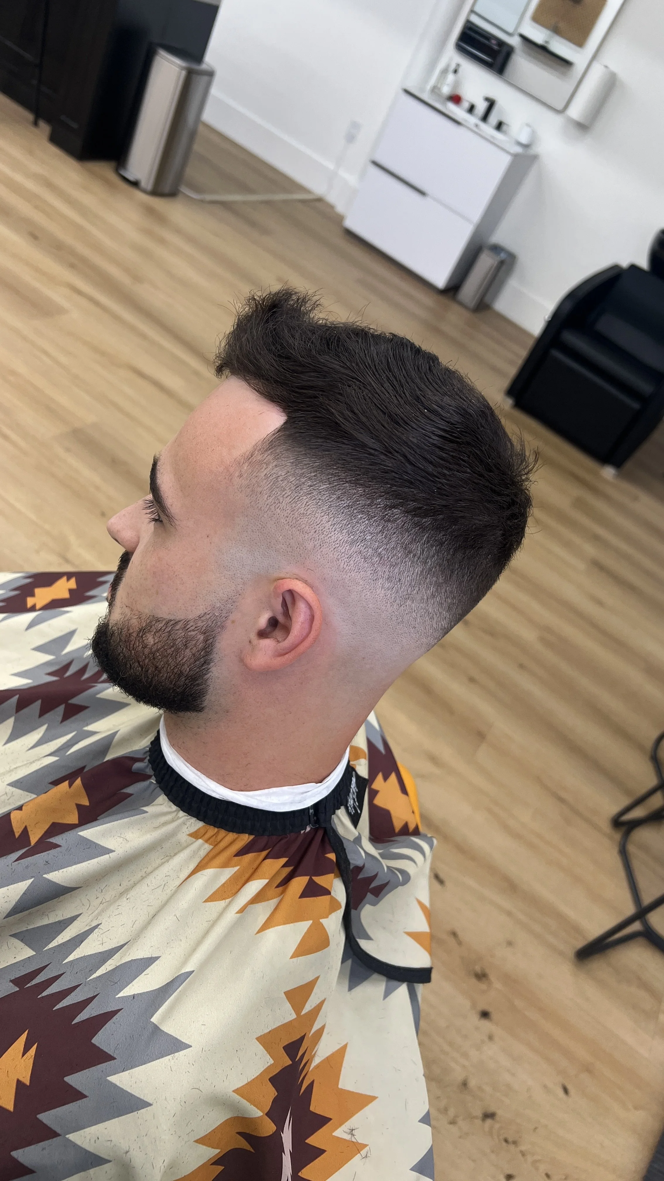 A man with dark hair in a modern haircut with short sides and longer top, a beard, and wearing a patterned cape in a barber shop, with wooden floors and barber stations in the background.