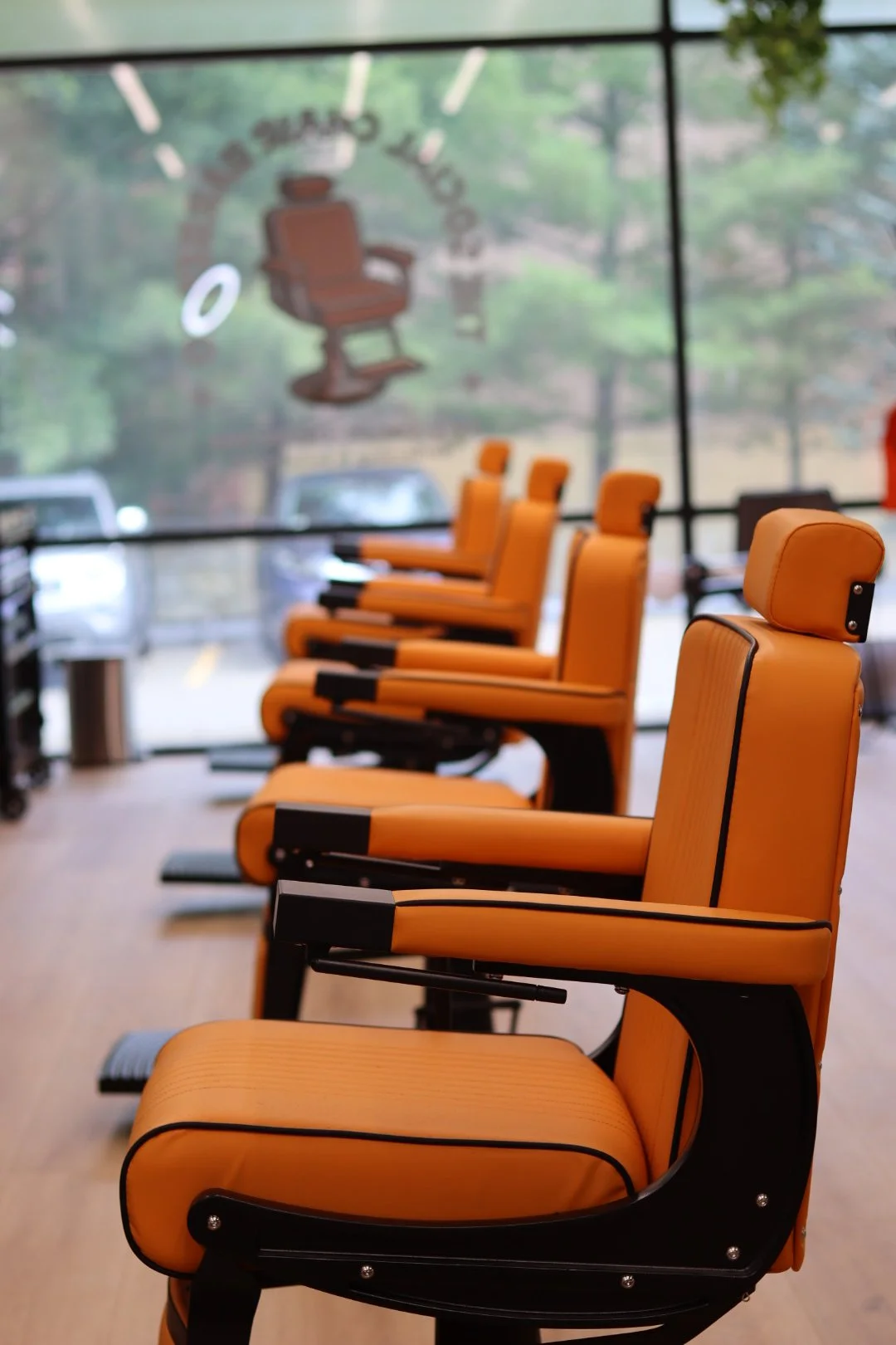 Row of orange barber chairs in a waiting area with a large window showing trees and parked cars outside. Perfect welcoming barbershop in Danvers Massachusetts 