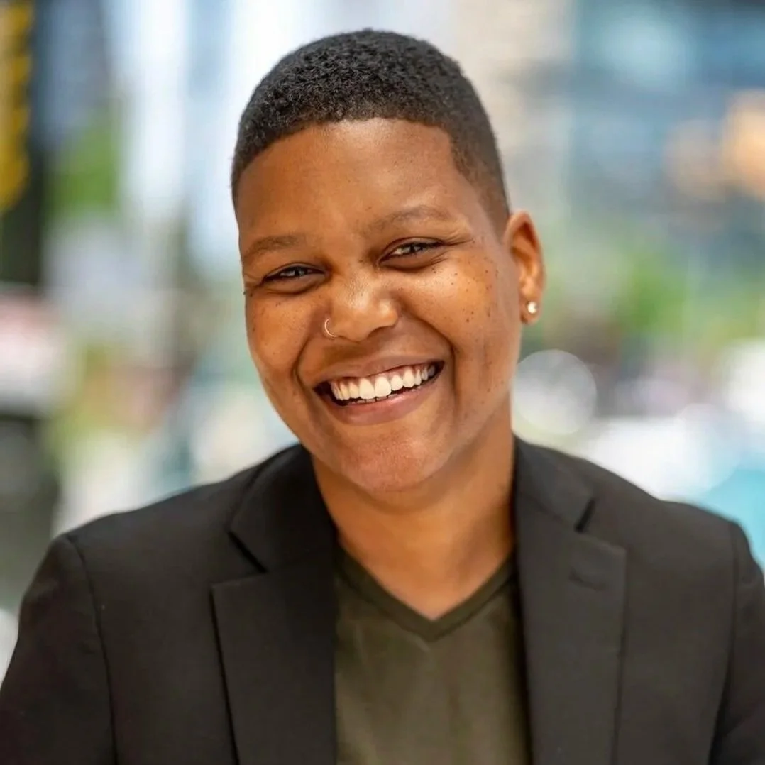A smiling woman with short hair, a nose ring, and earrings, wearing a black blazer over a dark shirt, in an indoor setting with blurred background.