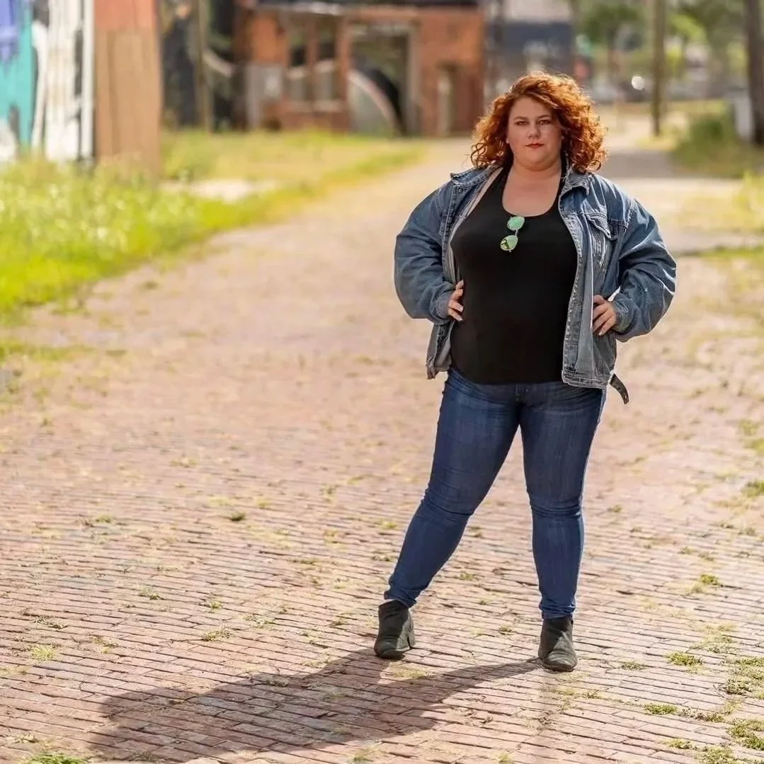 A woman with curly red hair standing confidently on a brick street with hands on her hips, wearing a black shirt, blue jeans, black shoes, and a denim jacket, in an outdoor urban setting.
