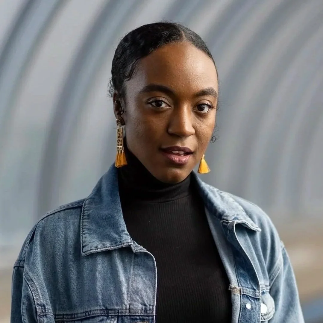 A young woman with dark hair in braids, wearing a black turtleneck, denim jacket, and yellow tassel earrings, standing against a blurred tunnel background.