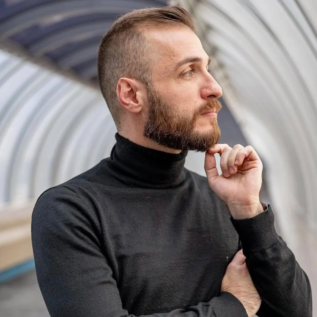 A man with a beard and short hair, wearing a black turtleneck, thoughtfully looking to the side with his hand resting on his chin inside a greenhouse or tunnel.