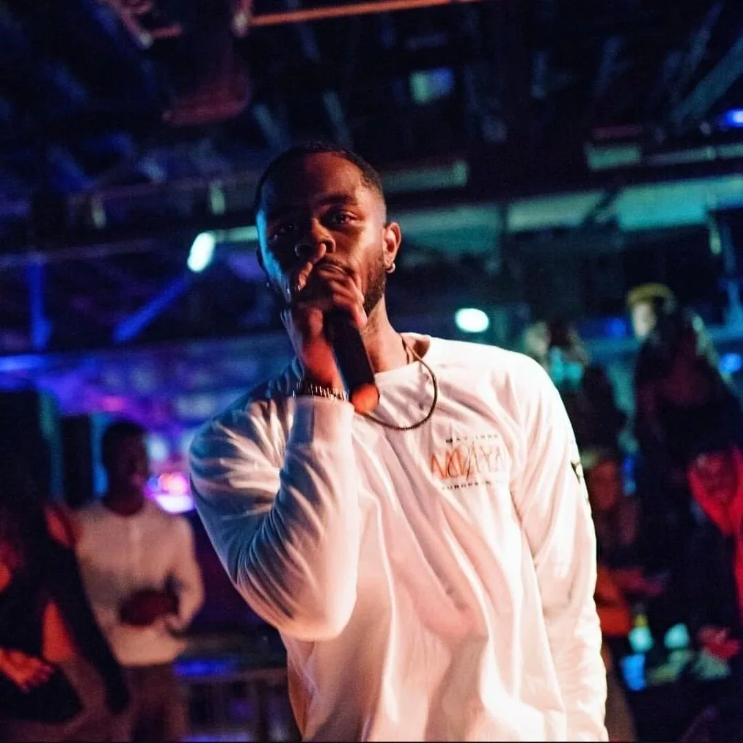A man with a beard and earring holds a microphone to his mouth in a dimly lit nightclub or concert setting, with colorful lights and blurred people in the background.