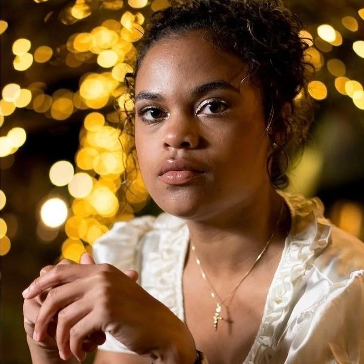 A young woman with dark curly hair and a serious expression, wearing a white top and gold necklace, is sitting outdoors at night with blurred warm yellow string lights in the background.
