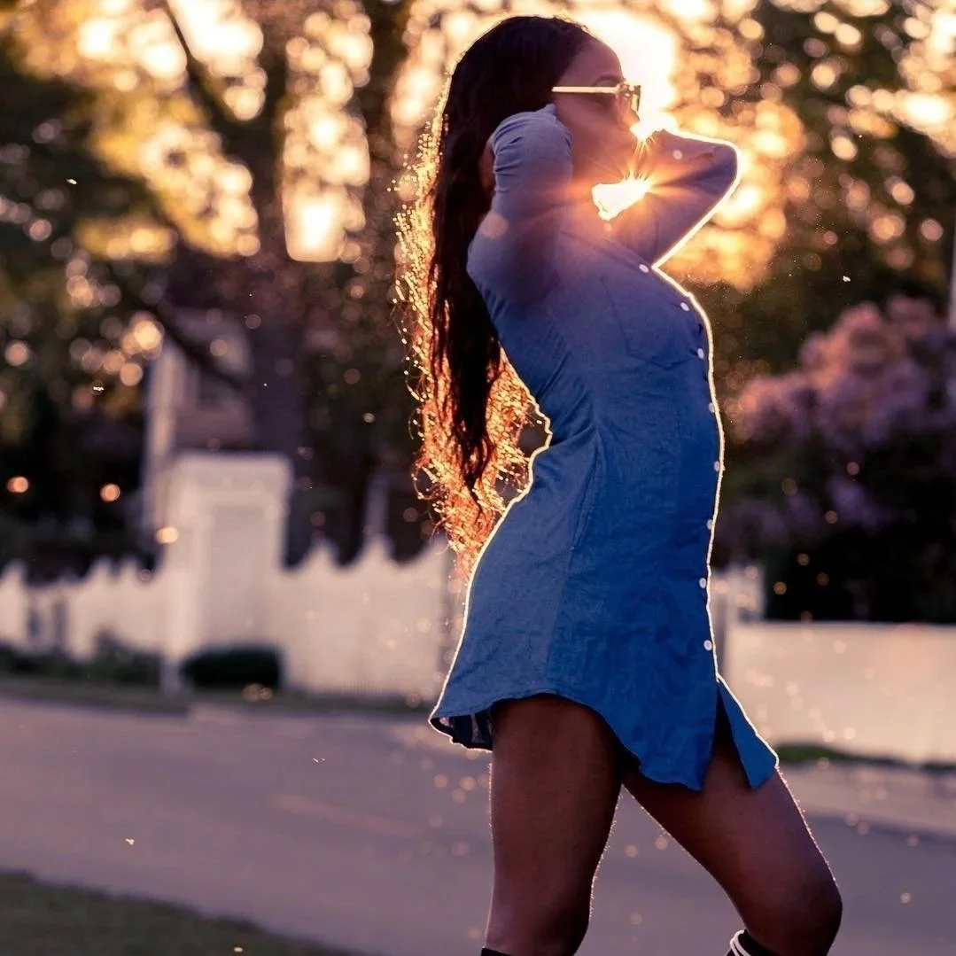 A woman in a blue dress and sunglasses standing outdoors during sunset, with sunlight illuminating her hair.