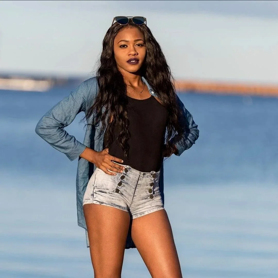 A woman with long curly black hair and dark lipstick standing outdoors by a body of water, wearing sunglasses on her head, a denim shirt, a black top, and high-waisted shorts.