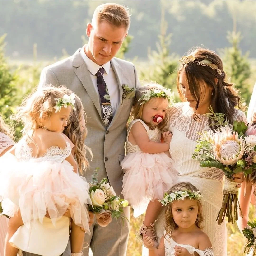 A wedding scene with a woman holding a bouquet of flowers, a man in a suit, and four young girls dressed in pink and white with flower crowns. The woman is gently holding one of the girls, and they are outdoors in a lush, green setting.