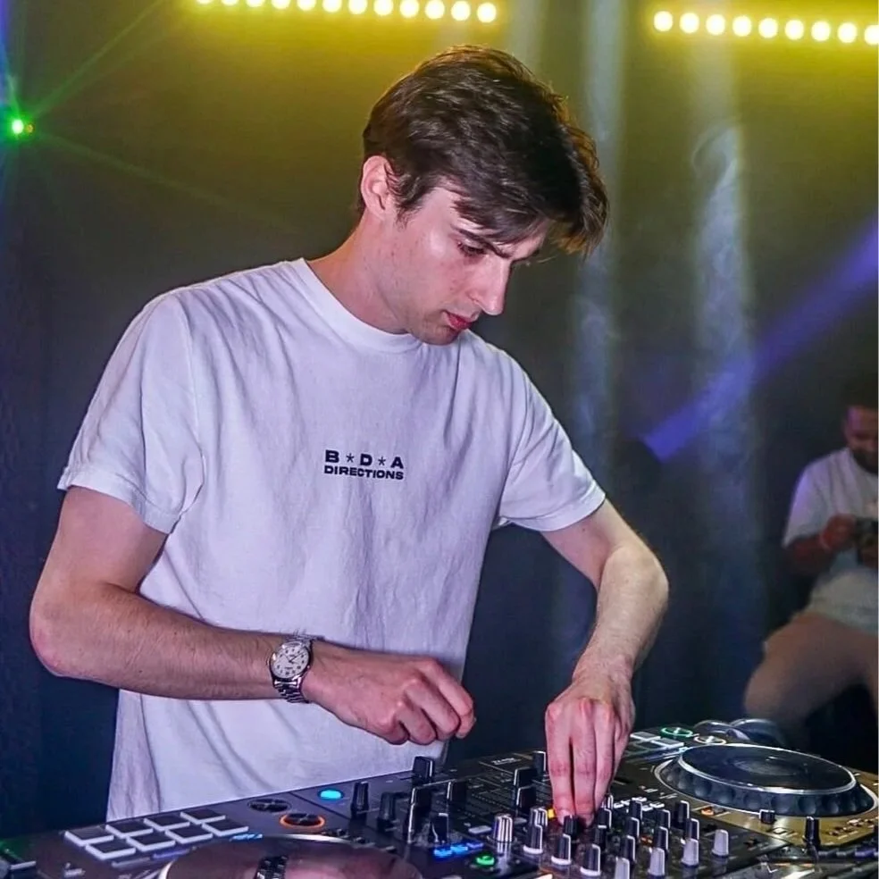 A young man with dark hair wearing a white t-shirt and a watch is DJing at a club or party, surrounded by colorful laser or stage lights. In the background, another person is seated, looking at their phone.